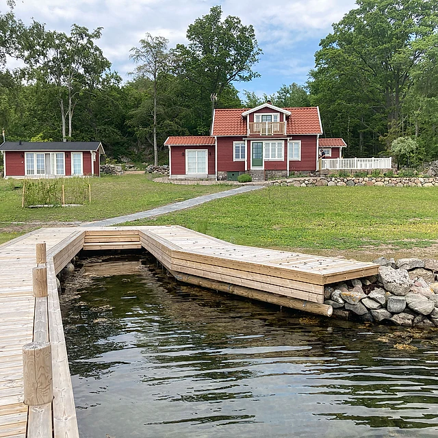 Brygga i ljust trä, vatten, grön gräsmatta, grusgång som leder upp till ett rött hus